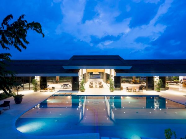 A modern building with a spacious outdoor pool is illuminated at night, surrounded by seating and greenery under a partly cloudy sky.