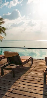 A serene poolside scene with lounge chairs, umbrellas, and palm trees overlooking a calm ocean under a bright sky.
