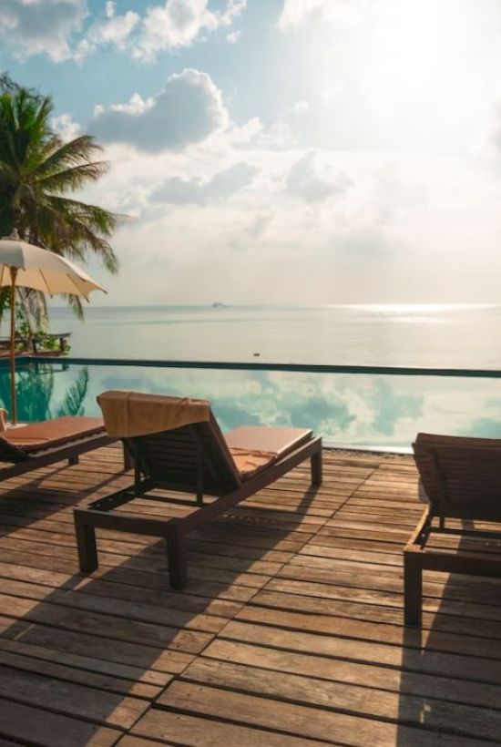 Sunlit poolside with lounge chairs and umbrellas overlooking a serene ocean view, surrounded by palm trees and a clear sky.