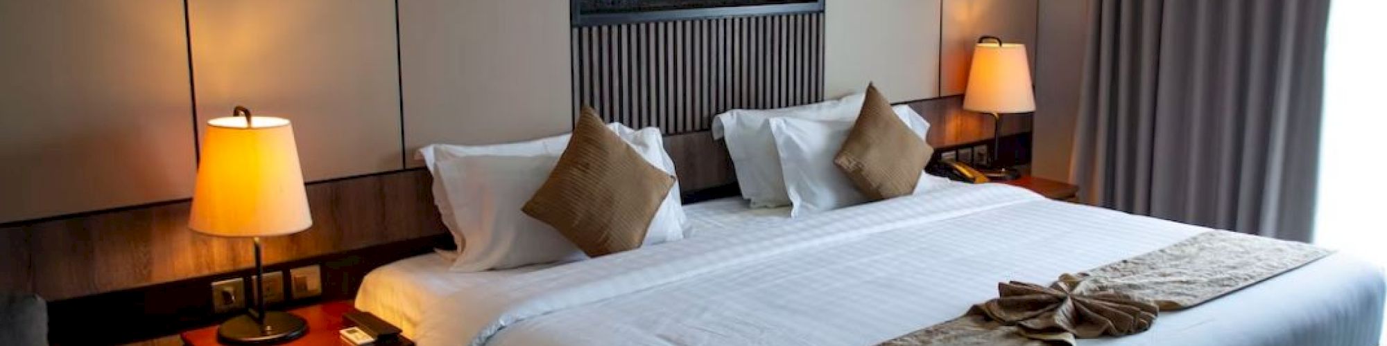 A cozy hotel room with a large bed, two bedside lamps, decorative cushions, art on the wall, and curtains covering a window.