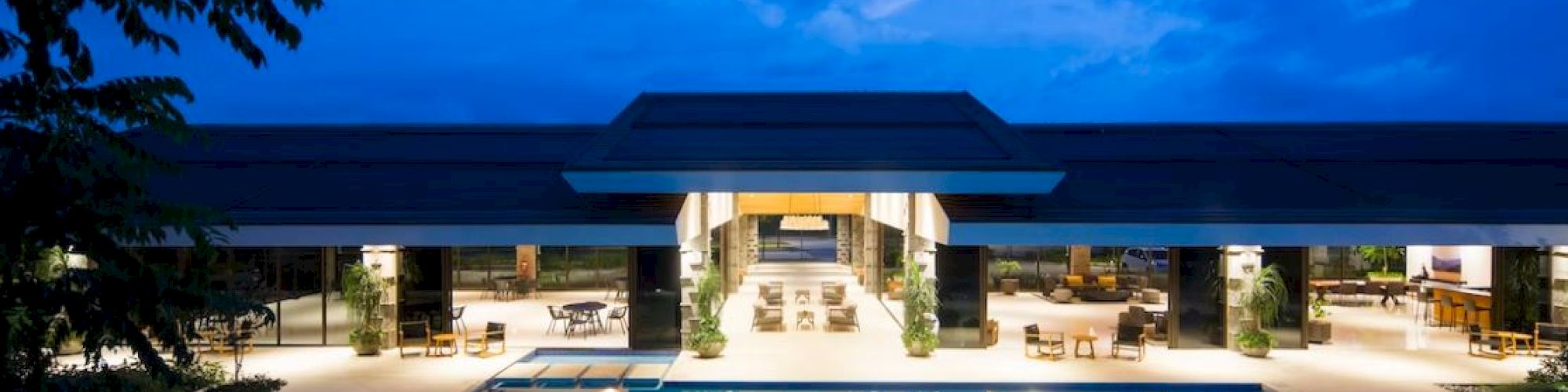 A modern building with large open spaces is reflected in a calm pool under an evening sky with scattered clouds.