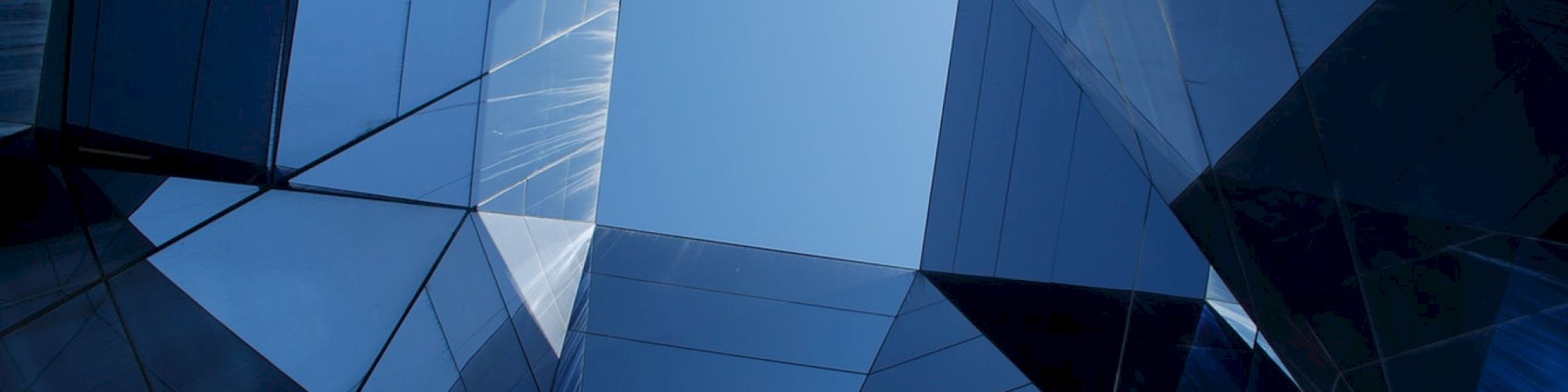 Geometric, reflective glass structures form a polygonal space, revealing a clear blue sky above within angular shapes.
