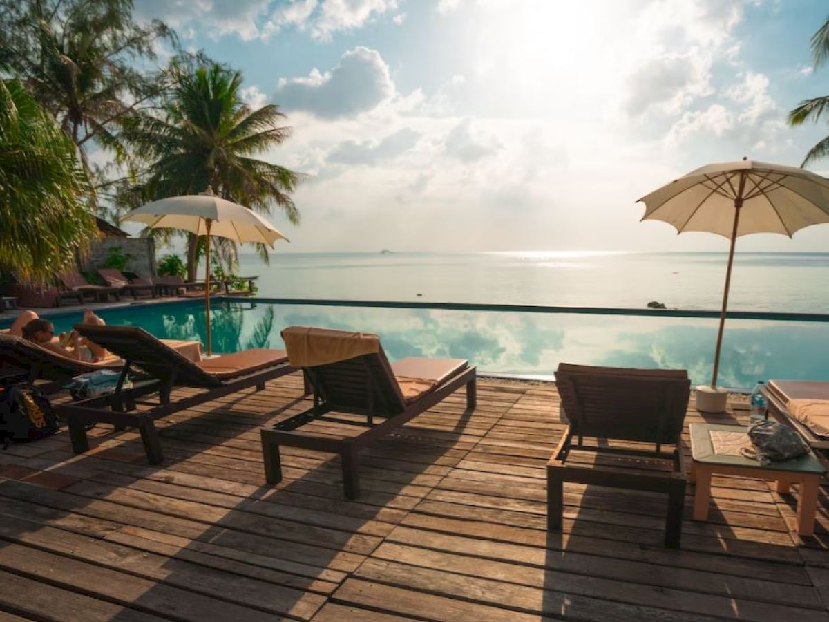A serene poolside view with sun loungers, umbrellas, palm trees, and a calm sea under a bright sky with scattered clouds.