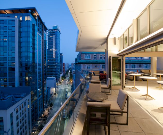 A cityscape at dusk showing a modern balcony with tables and chairs overlooking a street lined with tall buildings.