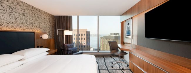 A contemporary hotel room features a large bed, modern decor, a wall-mounted TV, and a workspace near a window with a city view.