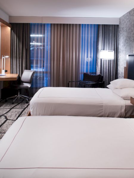 The image shows a modern hotel room with two beds, a TV, a desk, and stylish decor featuring patterned carpet and wallpaper.