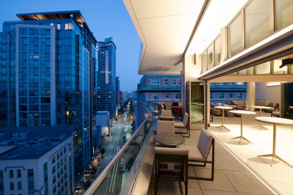 A modern rooftop terrace with tables and chairs overlooks a cityscape with tall buildings at twilight, featuring a glass railing.