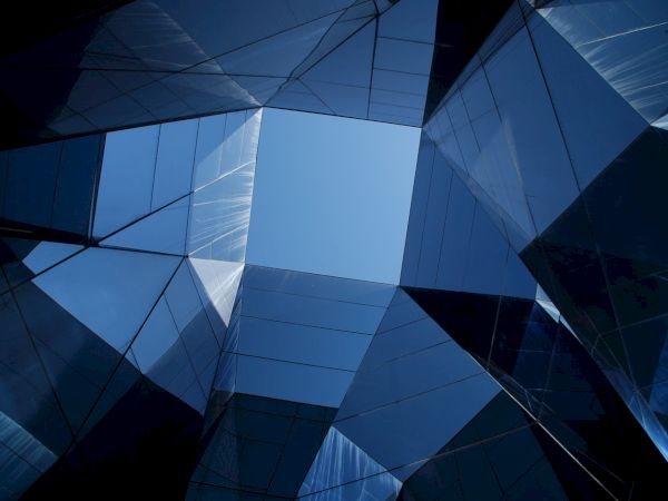 The image shows a geometric architectural space with reflective glass surfaces creating a kaleidoscopic view towards a clear blue sky.