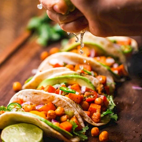 Tacos filled with chickpeas, tomatoes, avocado, and cilantro on a wooden board, with lime being squeezed over them.