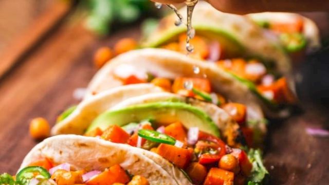 Tacos filled with chickpeas, tomatoes, avocado, and cilantro on a wooden board, with lime being squeezed over them.