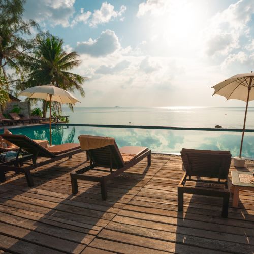 Poolside view with lounge chairs, umbrellas, and lush trees overlooking the ocean at sunrise or sunset, creating a tranquil atmosphere.