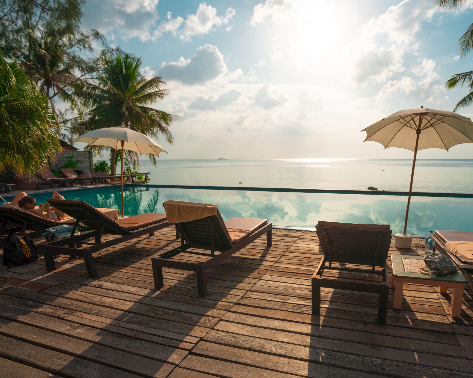 Lounge chairs and umbrellas by a pool with a view of the ocean, set under a cloudy sky and surrounded by palm trees.