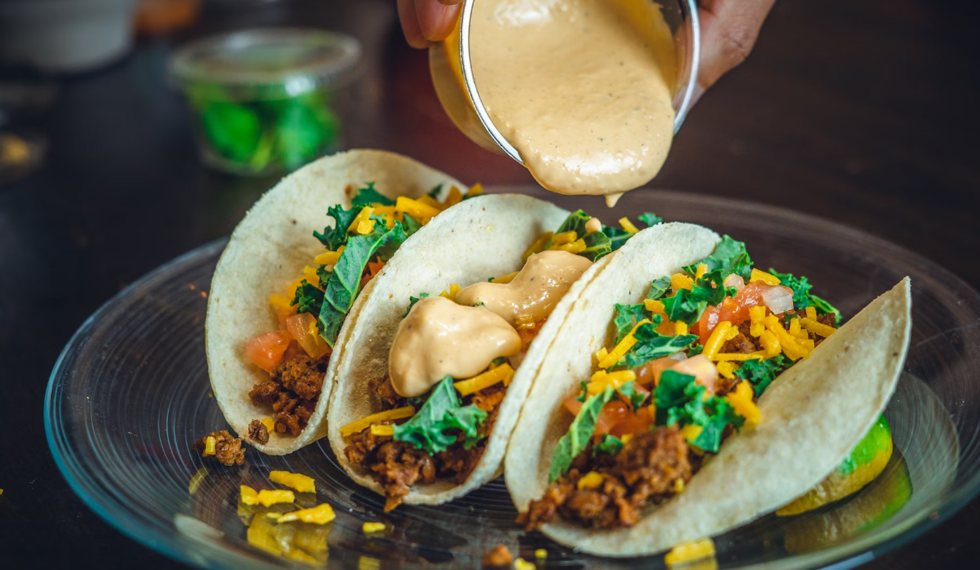 Three tacos on a glass plate are being topped with a creamy sauce, containing ingredients like lettuce, cheese, and tomatoes.