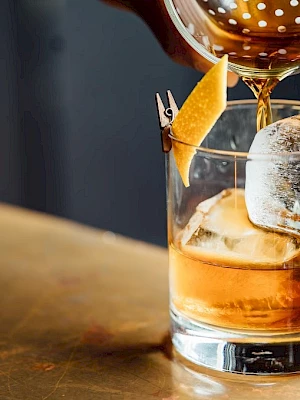 A hand pours a cocktail into a glass with ice and a lemon peel garnish, on a wooden surface.
