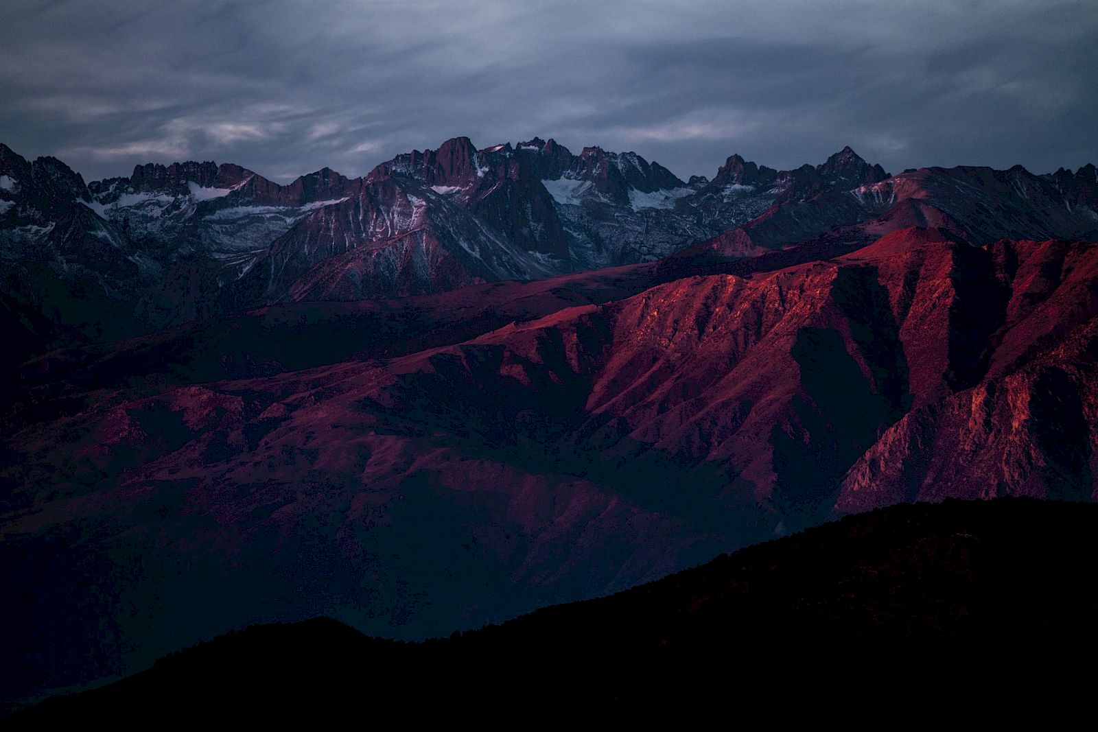 A mountain range bathed in red light under a cloudy sky at sunset.