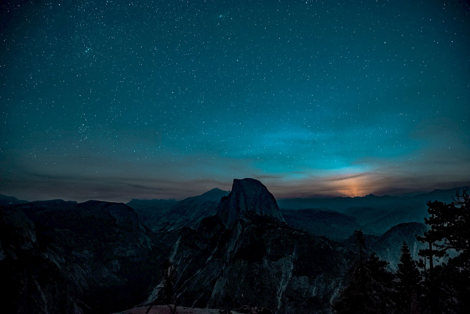 A starry night sky over a mountain silhouette with trees in the foreground and a gentle glow near the horizon.