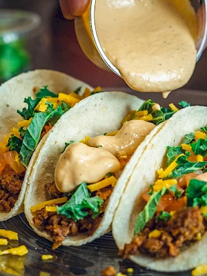 Three tacos loaded with ground meat, cheese, lettuce, and tomatoes have sauce being poured over them on a plate.