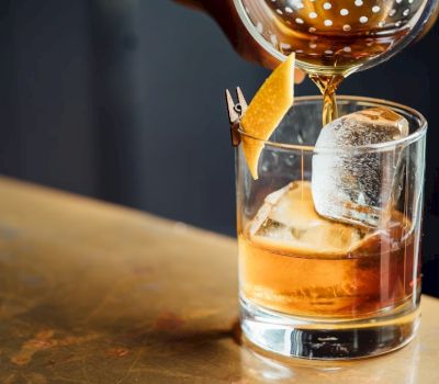 A cocktail is being prepared with a brown liquid, ice, and an orange peel.