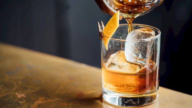 A drink is being poured into a glass with ice and a citrus twist garnish, on a bar top.