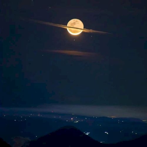 A bright full moon is aligned with a thin cloud, resembling Saturn with its rings, above a dark landscape with scattered lights.