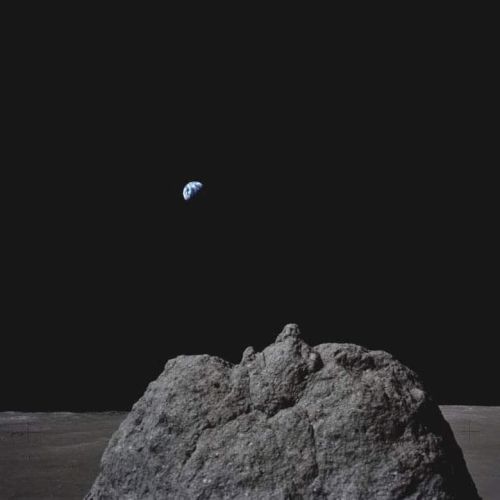A large rock is in the foreground on the lunar surface, with Earth visible in the distant black sky above it.