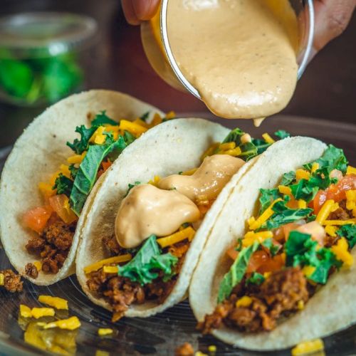 Three tacos on a plate filled with ground meat, shredded cheese, leafy greens, and diced tomatoes, being topped with creamy sauce.