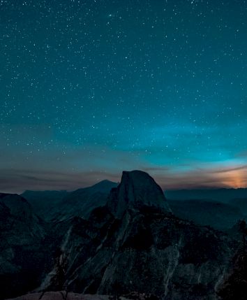A starry night sky over a mountainous landscape with silhouettes of trees, featuring a prominent peak in the foreground.