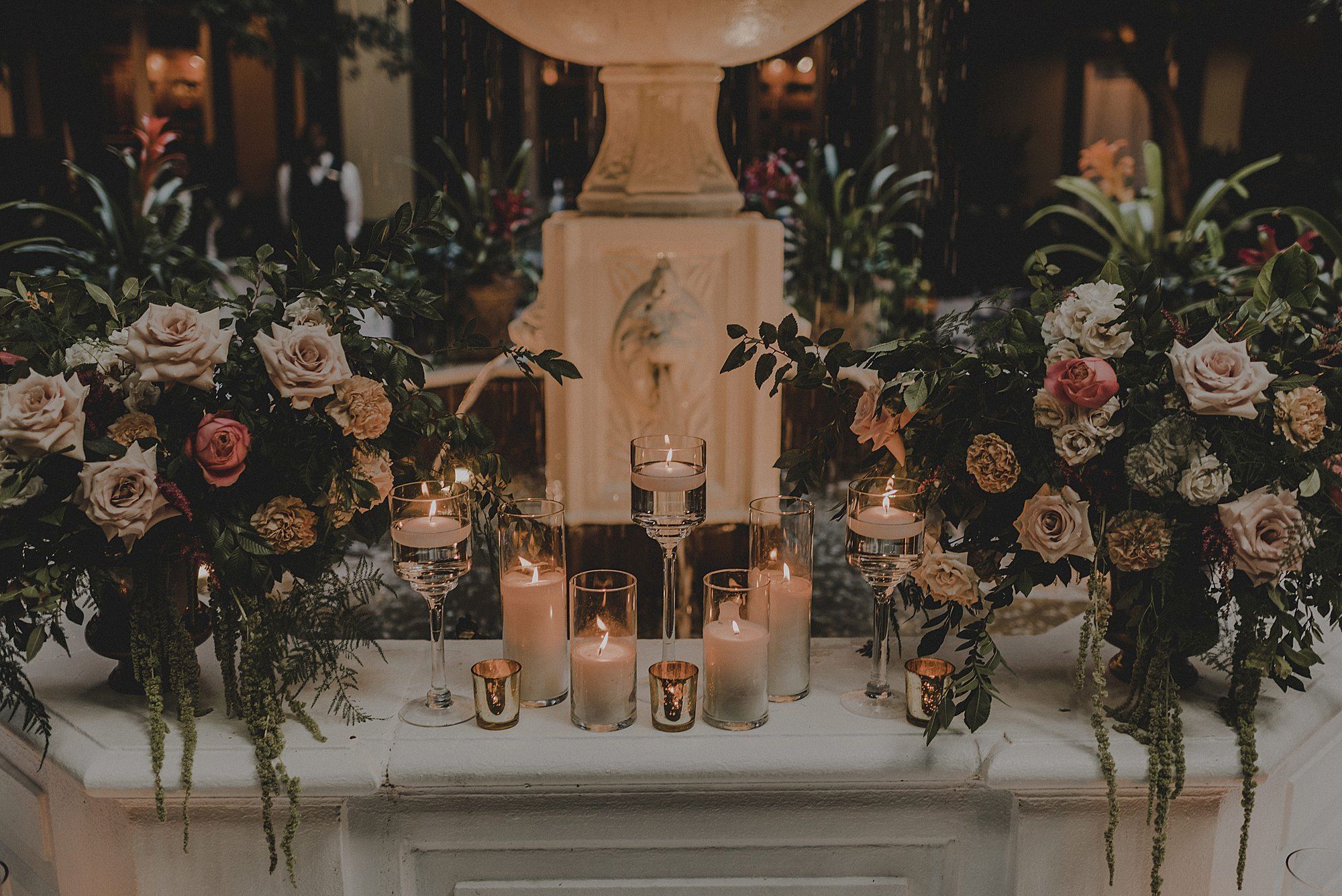 The image shows a decorative setup with lit candles, flower arrangements, and greenery in front of a stone fountain.