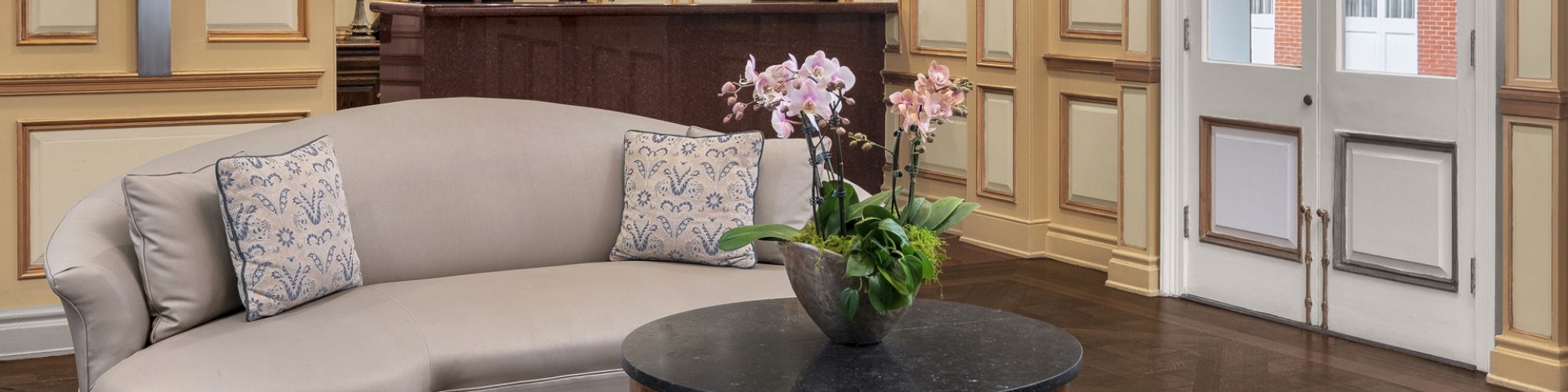 A stylish hotel lobby with a curved sofa, a round coffee table with orchids, and a reception desk in the background.