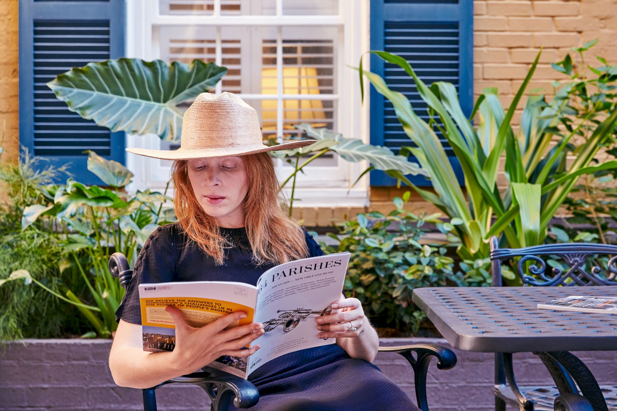 A person in a wide-brimmed hat sits outdoors, reading a magazine titled "64 Parishes," surrounded by lush greenery and a table.