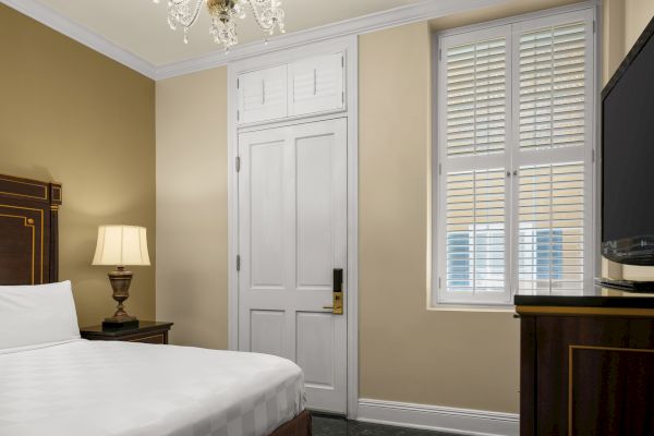A hotel room with a bed, wooden headboard, nightstand, lamp, TV, door, window with shutters, and chandelier on the ceiling.