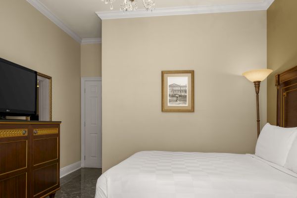 A hotel room with a bed, framed artwork, TV, and a floor lamp, decorated in neutral tones, offering a cozy atmosphere.