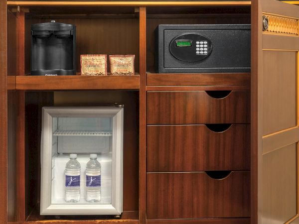 The image shows a wooden cabinet with a coffee maker, safe, small fridge with water bottles, and drawers.