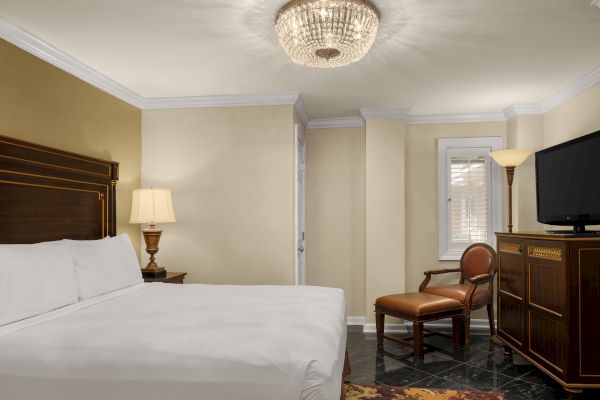 A hotel room with a large bed, ornate chandelier, wooden furniture, and a TV on a cabinet. There's a soft lamp near the bed.