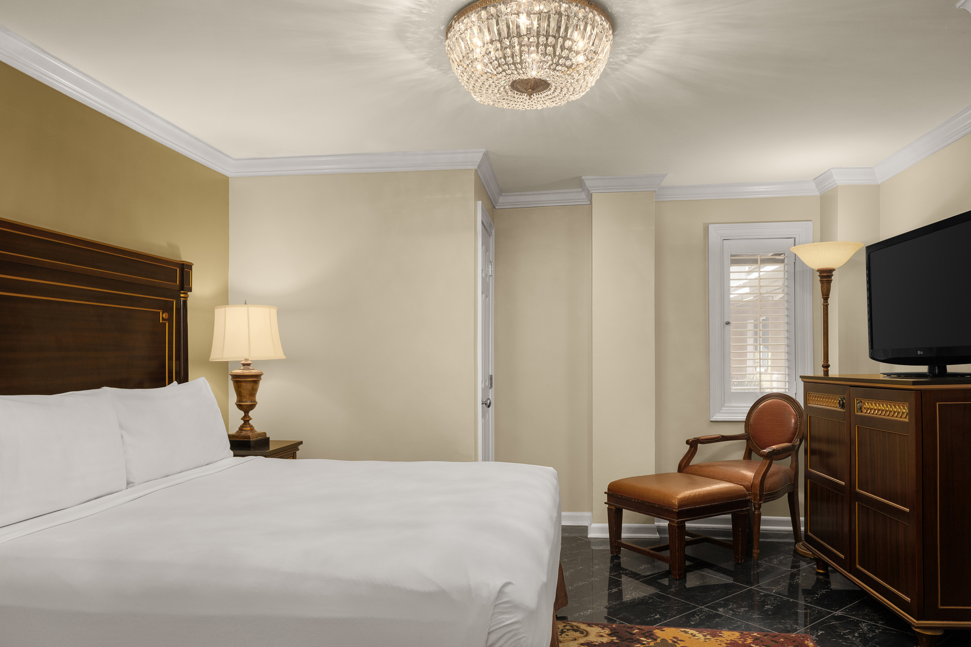 A hotel room with a king-sized bed, a chandelier, a TV on a dresser, and a chair.
