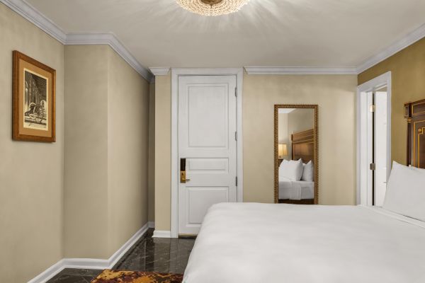 A hotel room features a bed, mirror, artwork, and a white door with a modern light fixture on the ceiling.