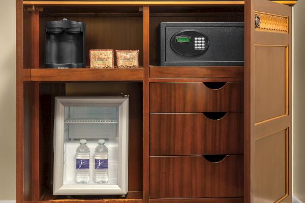 A hotel cabinet with a coffee maker, coffee packs, digital safe, mini fridge with two water bottles, and drawers.