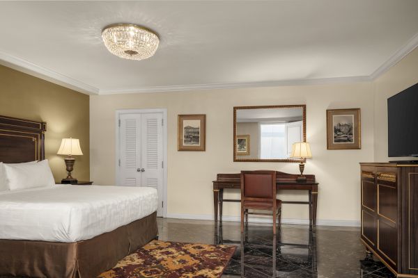 A neatly arranged hotel room with a bed, ornate desk and chair, large mirror, paintings, TV, and elegant lighting on the ceiling.