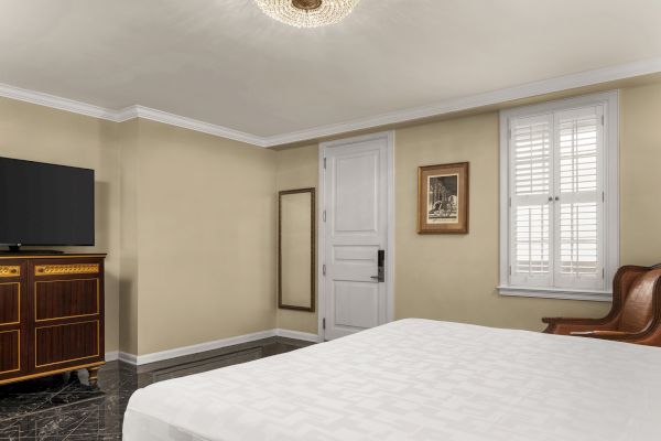 A room with a bed, wooden TV cabinet, mirror, chair, white door, framed picture, and window with shutters.