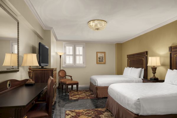 A hotel room with two beds, a desk, a chair, a TV, lamps, a window, and framed wall art, decorated in a classic style.