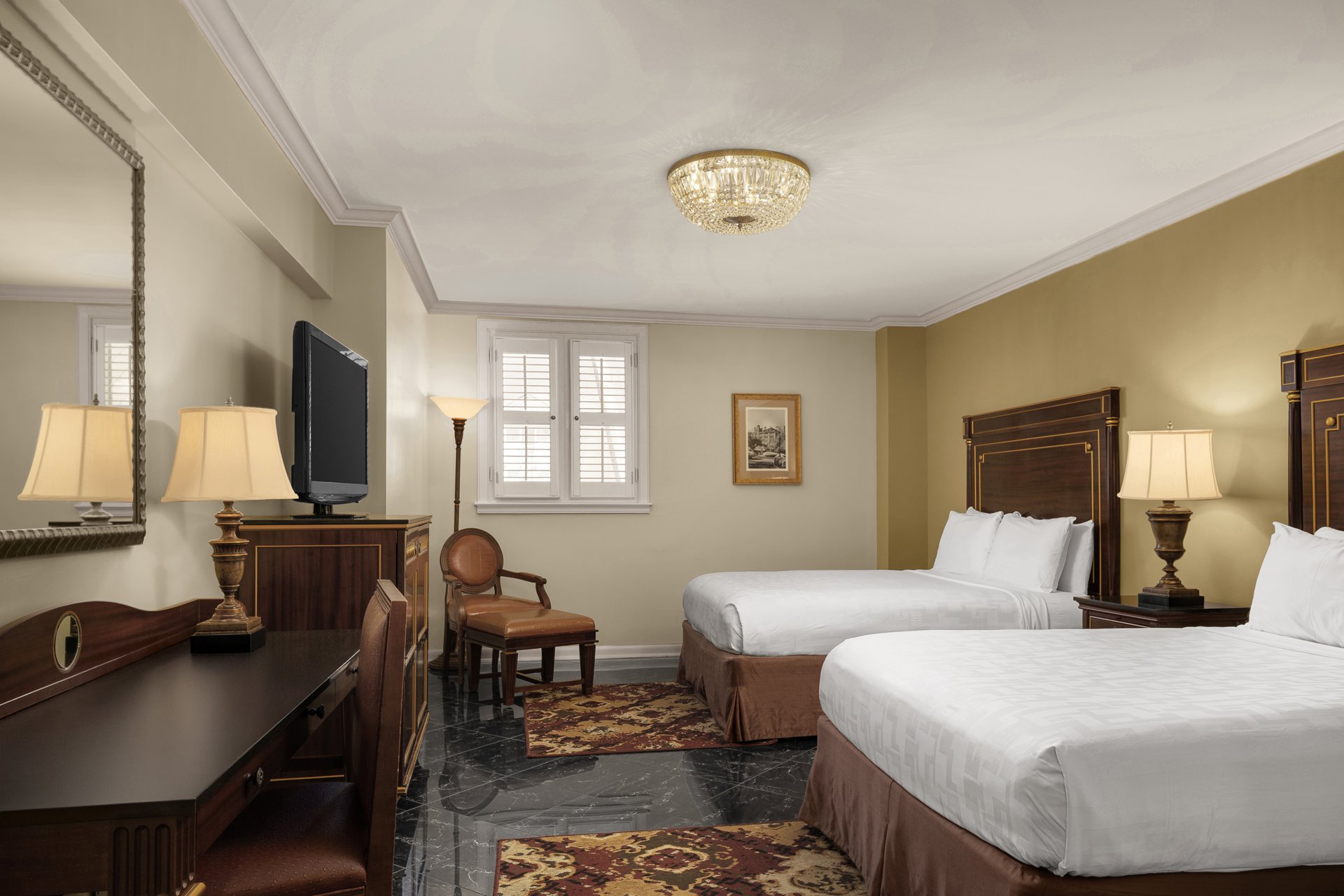 A hotel room with two beds, a desk, a TV, and a chandelier
