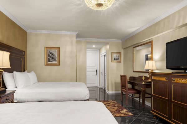 This image shows a hotel room with two beds, a desk, chair, TV, lamp, decorative mirror, and elegant lighting fixture on the ceiling.