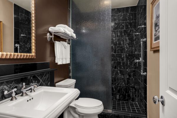 The image shows a modern bathroom with a white sink, a toilet, black marble shower, towels on a rack, and a framed picture on the wall.