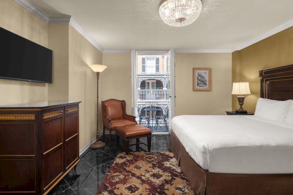 A hotel room with a bed, armchair, lamp, TV, and dresser; open doors lead to a balcony with a table and chairs.