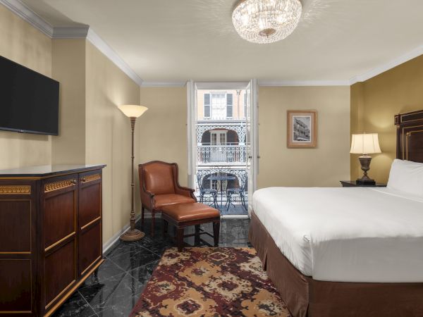 A hotel room with a bed, armchair, lamp, TV, and dresser; open doors lead to a balcony with a table and chairs.