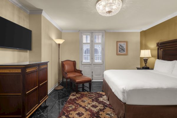 A cozy, elegant hotel room with a large bed, leather chair, artwork, TV, and ornate furniture, featuring warm lighting and decor.