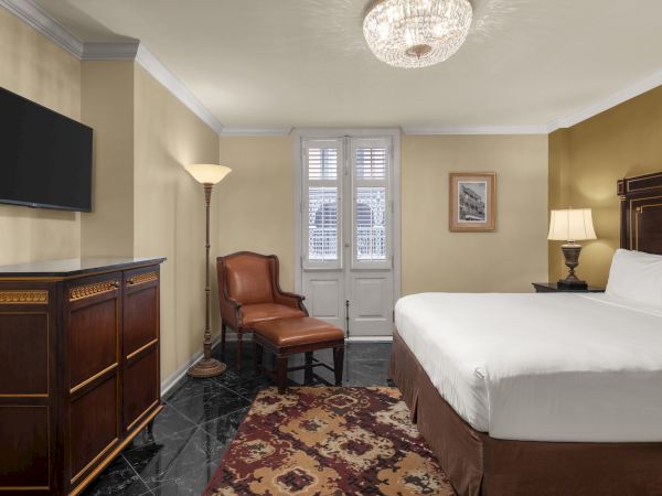 A cozy, elegant hotel room with a large bed, leather chair, artwork, TV, and ornate furniture, featuring warm lighting and decor.