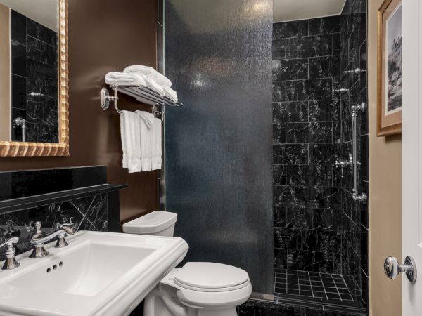 A modern bathroom with a white sink, toilet, and a black marble shower, featuring towels on a rack and a large framed mirror.