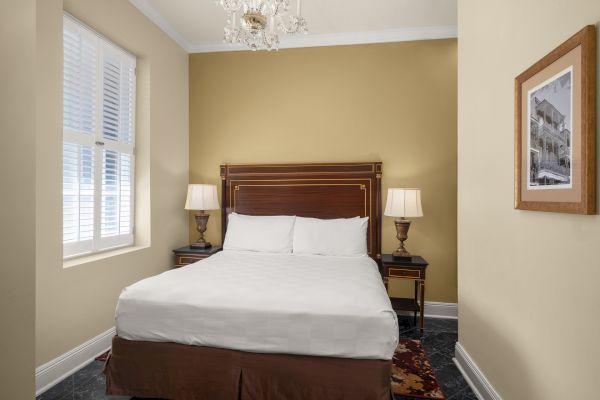 A cozy bedroom with a bed, two lamps on side tables, a window with shutters, and a framed picture on the wall completes the decor.