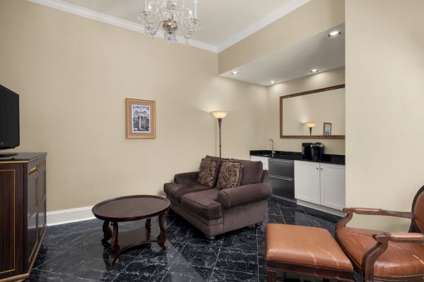 A living area with a brown sofa, armchair, and coffee table. There's a TV, floor lamp, chandelier, artwork, and a kitchenette with a mirror.